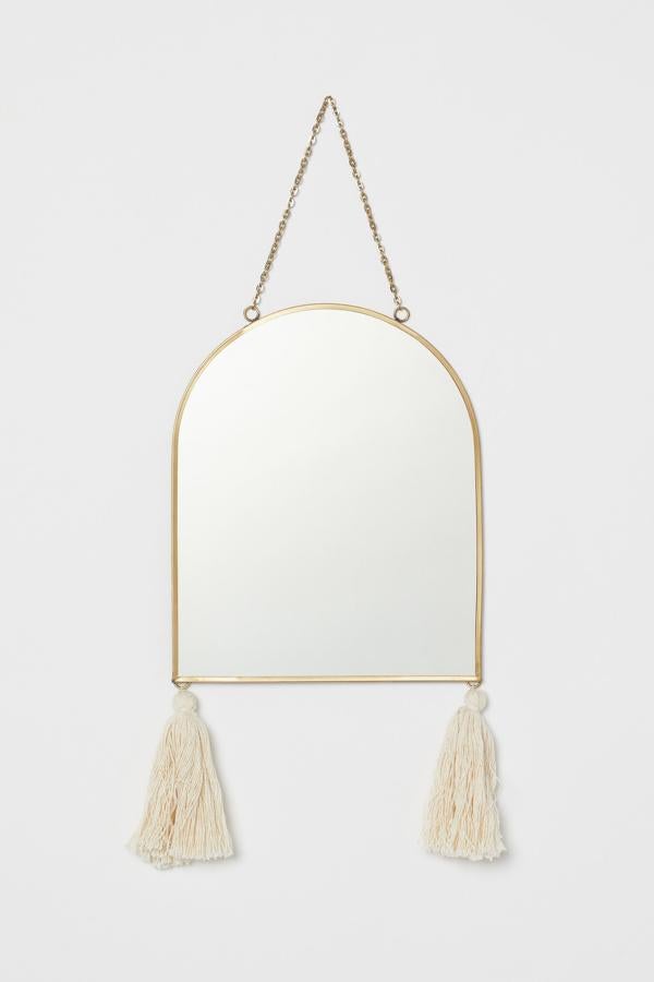 Este espejo con marco fino en metal dorado arqueado en la parte superior y dos borlas de tela en las esquinas inferiores tiene unas medidas de 20,5x25 centímetros y un precio de 15,99 euros.