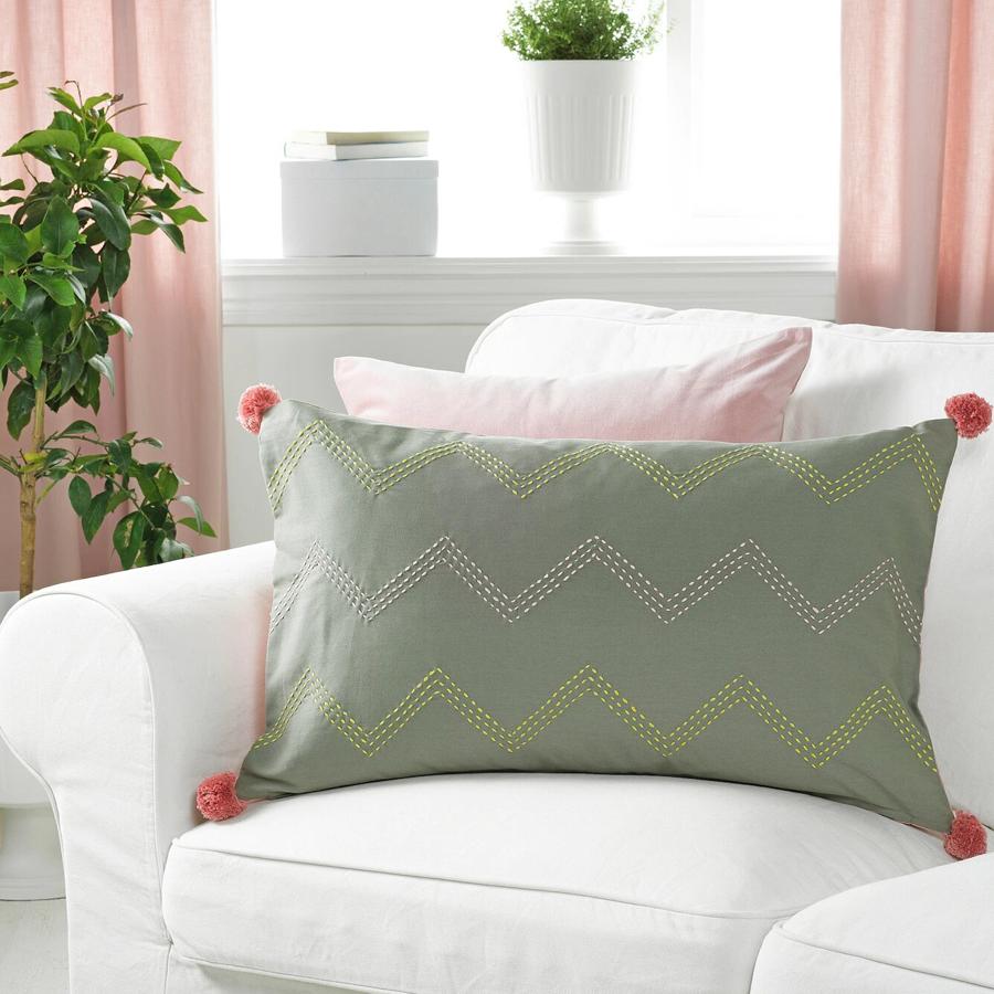 Esta funda de cojín confeccionada a mano forma parte de una iniciativa social. En color verde con pompones rosas, tiene unas medidas de 40x65 centímetros y un precio de 12 euros.