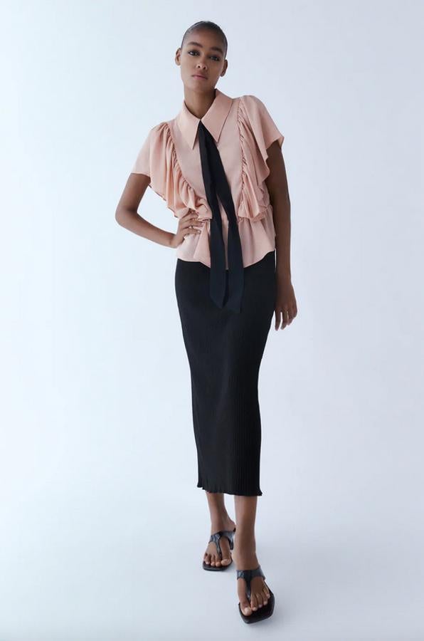 Con cuello solapa y manga corta, esta bonita blusa en rosa claro de Zara viene con cintura elástica, cierre frontal con botones, lazo combinado a contraste y volantes desde la parte frontal de la cintura hasta la espalda, cayendo sobre los hombros. Cuesta 22,95 euros y podemos encontrarla entre las tallas XS y XL y también en color blanco.