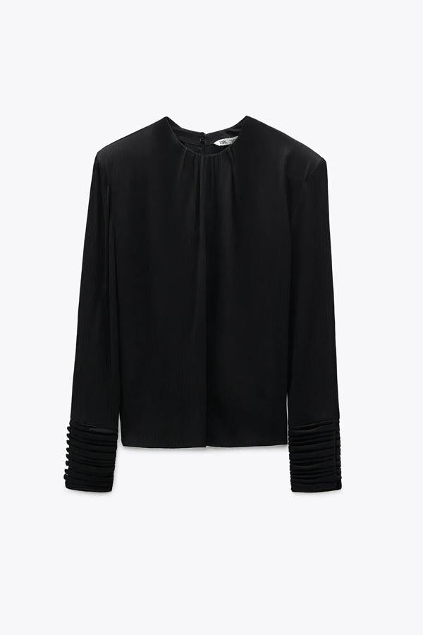 Esta blusa satinada con hombreras acabada en puño con botones forrados y pliegues cuenta con cierre en la espalda con abertura y botón. En color negro, es de Zara y tiene un precio de 29,95 euros.