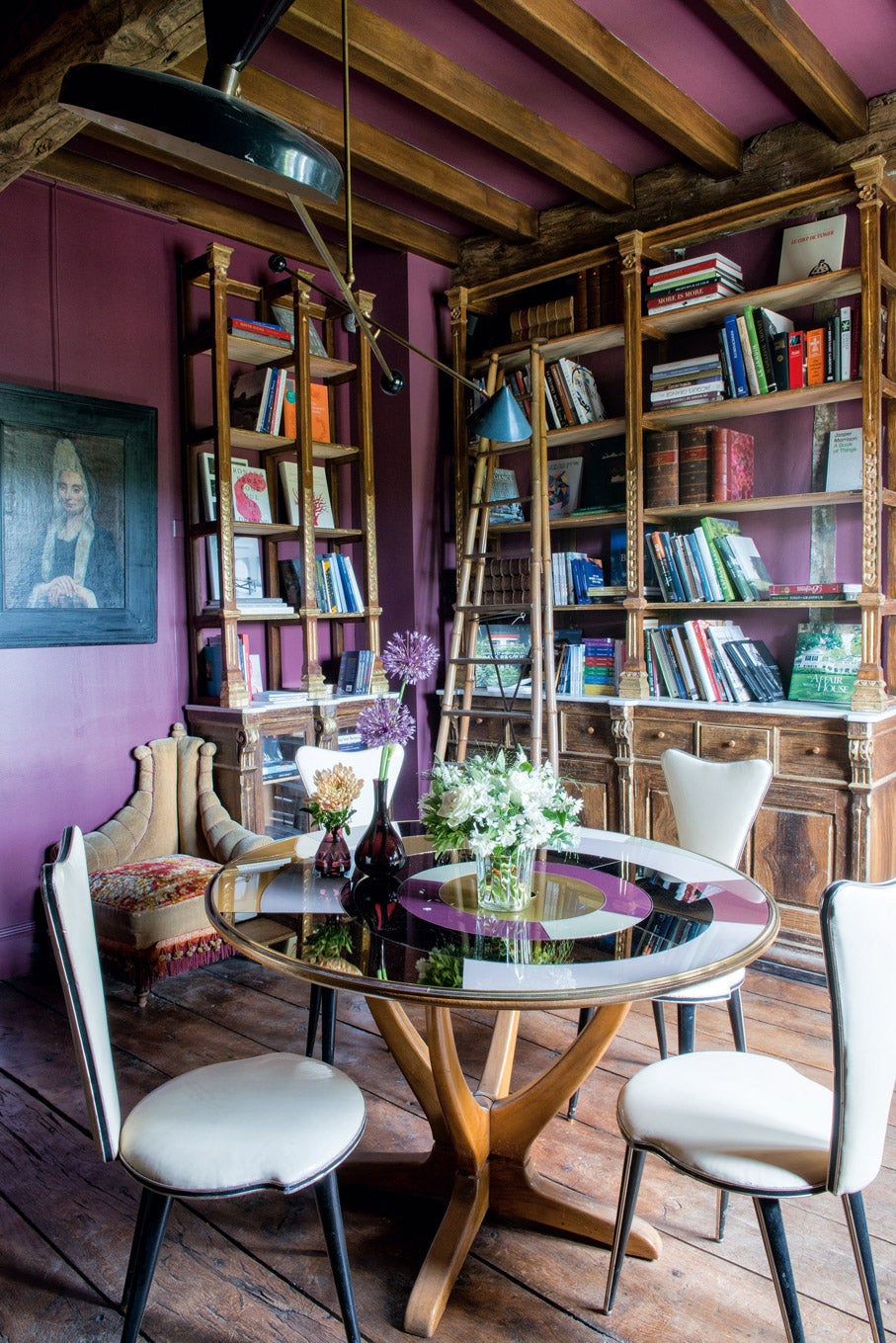 En la biblioteca, con las paredes en color morado, estantería del siglo XIX y mesa y sillas de los años 50. Detrás, butaca estilo Napoléon III con pasamanerías.