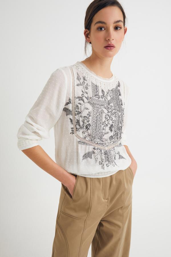 Esta blusa de plumeti en color crudo con bordado en negro en la parte delantera cuenta con manga larga y cuello a la caja y tiene un precio de 23,99 euros.