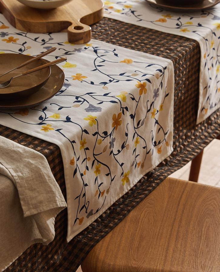 En color blanco y con estampado flora en azul y amarillo, este camino de mesa confeccionado en algodón satén tiene una medida de 50x160 centímetros y un precio de 25,99 euros.