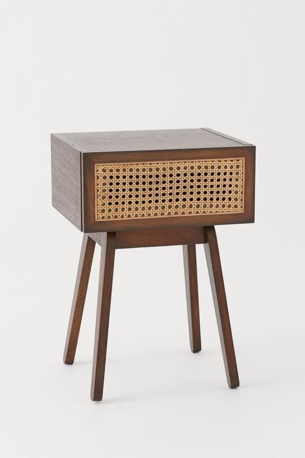 Disponible en marró o negro, esta mesita de noche en madera mercantil y cajón con frente de ratán tiene un precio de 129 euros.