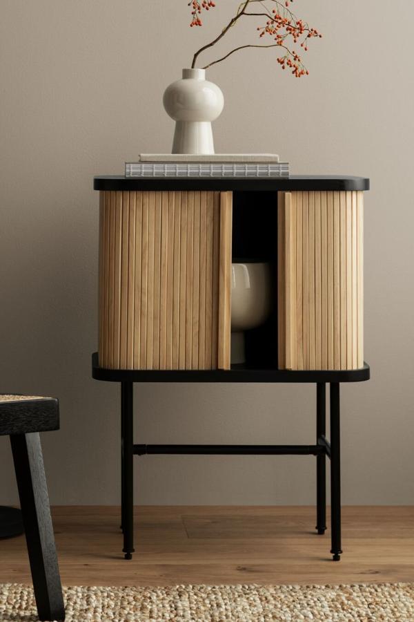 Esta mesita en madera MDF con chapa de madera de fresno cuenta con esquinas redondeadas y patas de metal pintado en negro. Tiene una altura de 46 centímetros, una altura de 60 y una profundidad de 27. Su precio es de 149 euros.