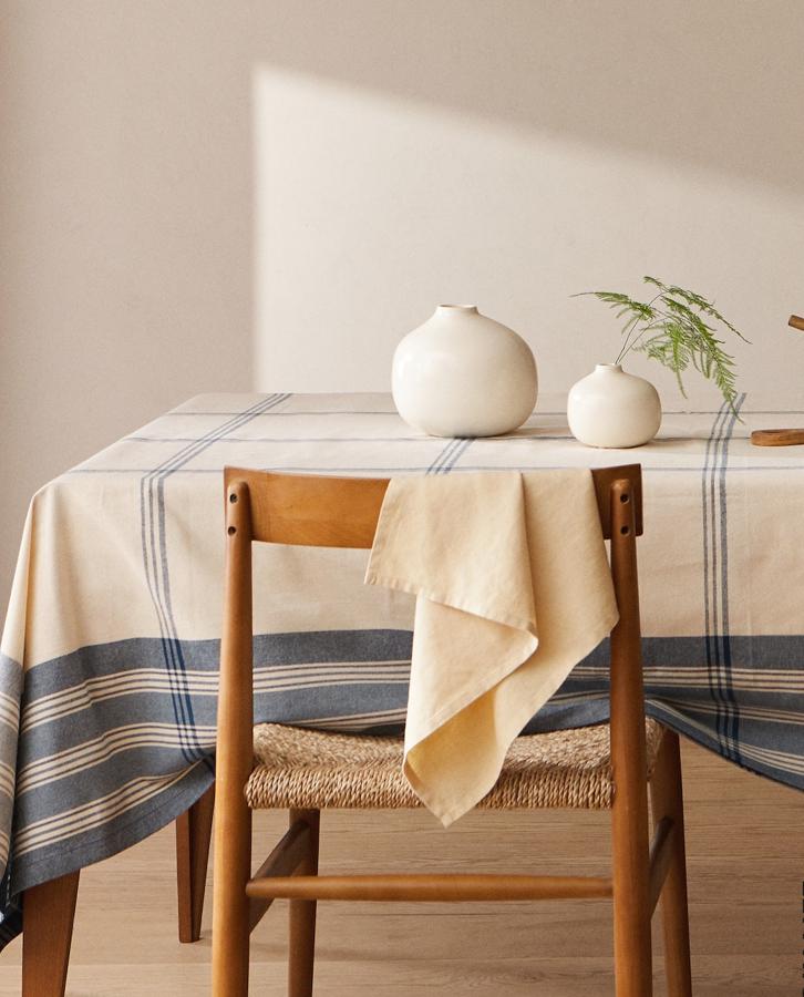 Para darle un aire fresco a la mesa, añade un mantel de rayas y también con pequeños toques azules. Este mantel es de algodón 100% orgánico y cuesta en Zara Home 25,99 euros.