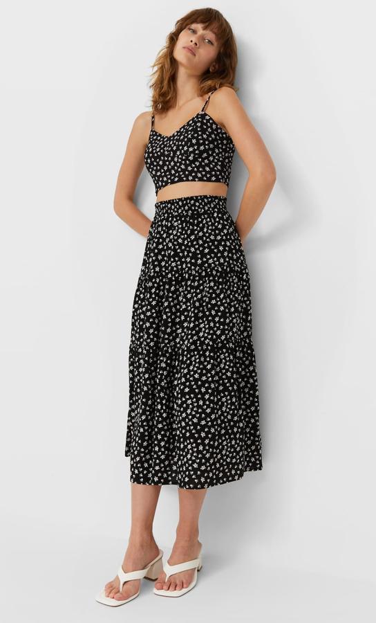 En color negro con pequeñas flores blancas, este conjunto está formado por una falda larga (19,99 euros) de tiro alto acabada en volantes y un top (12,99 euros) de tirantes con escote de pico y tirante fino regulable.