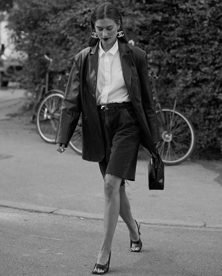 De estética ochentera. Esta 'influencer' combina chaqueta 'oversize' masculina de cuero, camisa abotonada hasta arriba, bermudas vaqueras en tono oscuro, pendientes maxi y tacones.