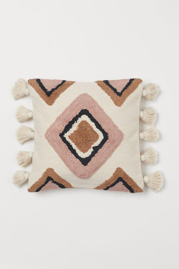 Esta funda de cojín beige con detalles en rosa y borlas grandes está disponible en varios colores. Con una medida de 50x50 centímetros y tejido jacquard tiene un precio de 24,99 euros.