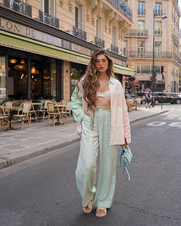 Un look en tonos pastel con pantalón ancho de cintura alta en verde claro, camisa oversize desteñida, bralette con aros, sandalias de velcro crema, cinturón de cadena dorado y bolso celeste. 