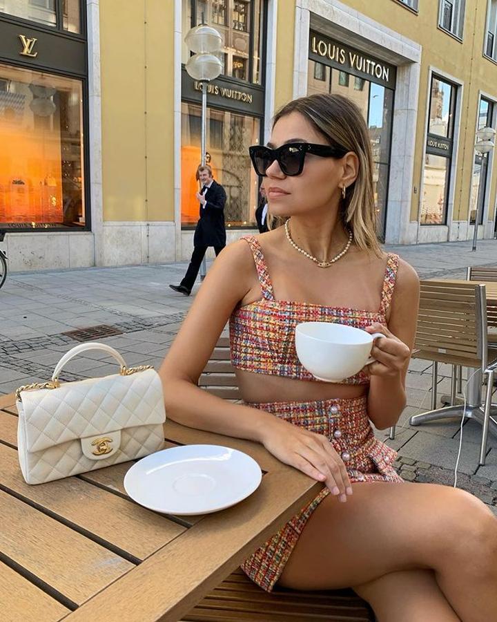 Un dos piezas de tweed multicolor con minifalda adornada por botones de perlas y bralette de tirantes anchos. Combina con joyería de perlas dorada y bolso acolchado, en tono crema.
