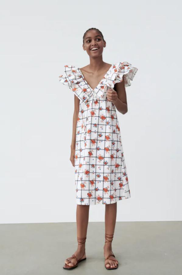 En color blanco, con estampado de cuadros a contraste en en negro y de flores en naranja, este vestido con escote de pico y manga sisa tiene detalle de volantes en el mismo tejido a lo largo del hombro y lazada en la pechera. Podemos comprarlo por 25,95 euros en las tallas S, M y L.