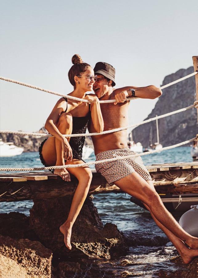 Gara lleva vestido de guipur negro sobre top y culot, todo de Givenchy; gafas de sol y pendientes de oro rosa y cerámica, ambos de Bvlgari. Pepe, bañador de Emidio Tucci, gorro bucket de Prada y reloj de Bvlgari.