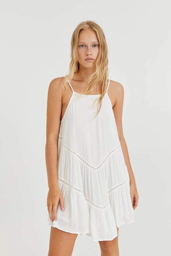 Este vestido corto de plumeti de color blanco cuenta con tirantes finos y escote recto. Con detalle de calados en diagonal y volantes, tiene un precio de 22,99 euros.