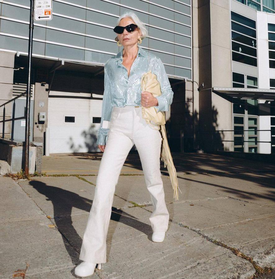 Las tendencias setenteras son lo más ahora. Grece apuesta por un estilismo disco que conjunta camisa brillante, pantalones de campana en blanco, botas de tacón con plataforma en el mismo tono y clutch crema con detalles de flecos.