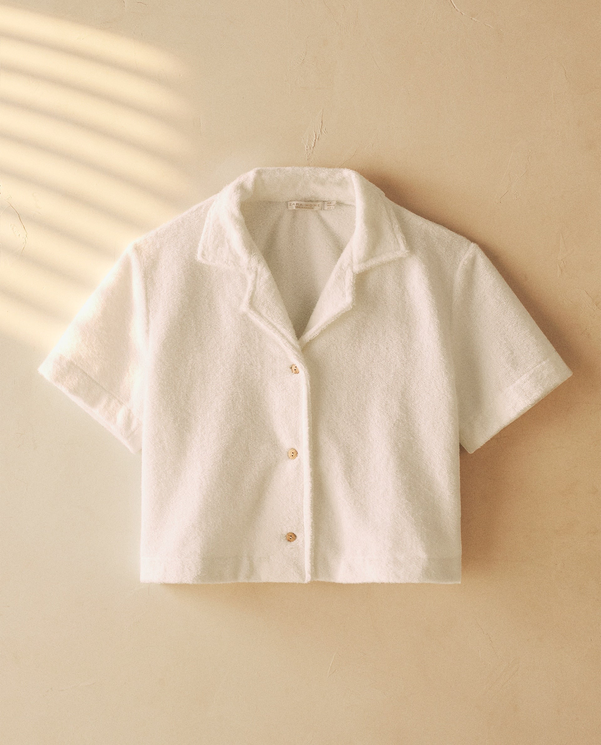 La estética setentera se cuela en nuestra selección con esta camisa blanca de silueta 'cropped' en tejido rizado y bambú, perfecta para llevar con unos vaqueros flare de cintura marcada. Cuesta 25.99 euros en Zara Home.
