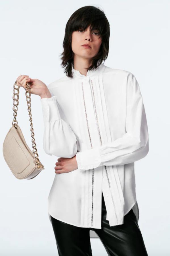 De cuello subido acabado en volante y mangas largas, esta camisa blanca de popelín es de Zara y viene con detalle de entredoses y jaretas en el frontal. Con cierre de botones ocultos por tapeta, cuesta 25,95 euros y está disponible entre las tallas XS y XL. 