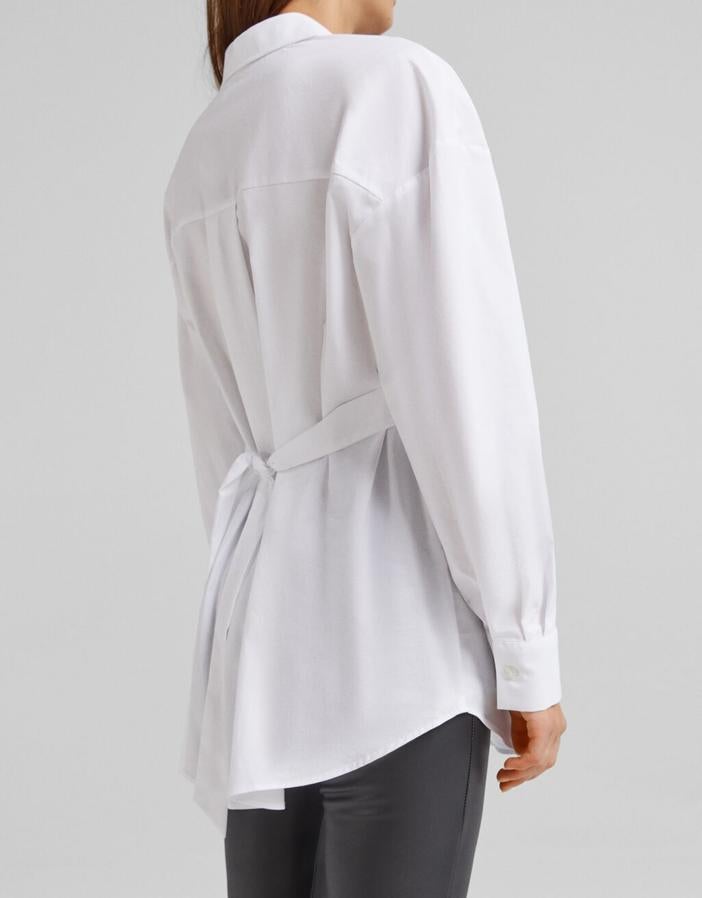Con cinturón a tono en la espalda que entalla la prenda, esta camisa blanca de Bershka con cuello de solapa y cierre frontal de botones cuesta 22,99 euros y está disponible entre las tallas XS y L. 