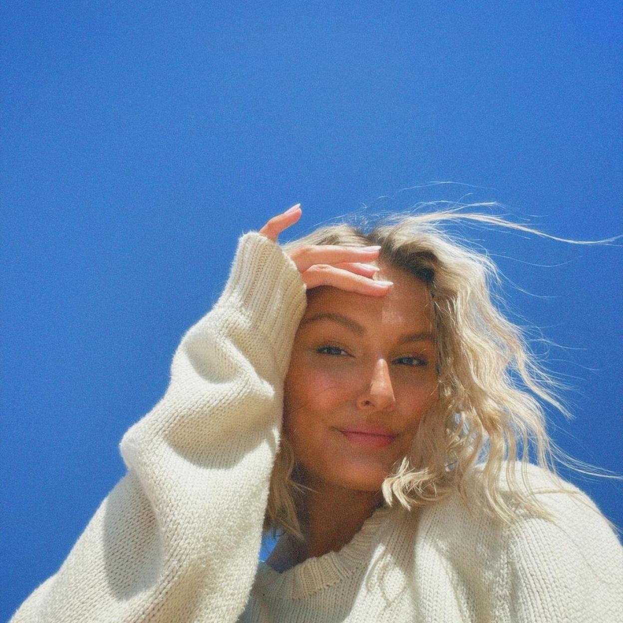 La influencer con el pelo ondulado y un jersey blanco. Su piel es lisa y sin imperfecciones. 