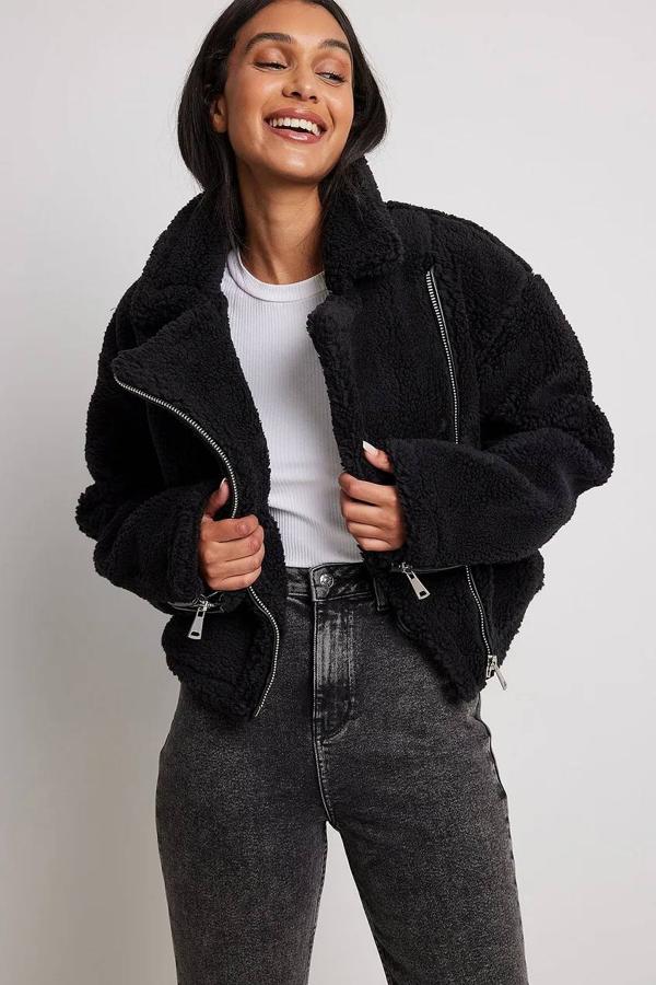 Chaqueta corta tipo biker en tejido de borreguillo con cuello solapa con muesca amplio, silueta relajada, manga larga y cierre cruzado mediante cremallera, en negro, de NA-KD. Puedes hacerte con ella por 80,95 euros. Cómpralo aquí
