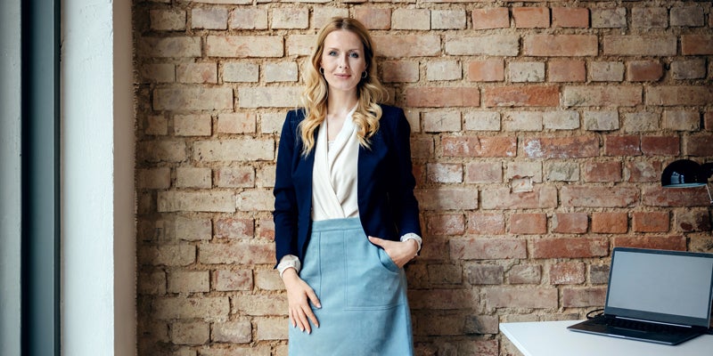 A portrait of a glamorous and small but successful business manager standing by a brick wall in her modern office space.