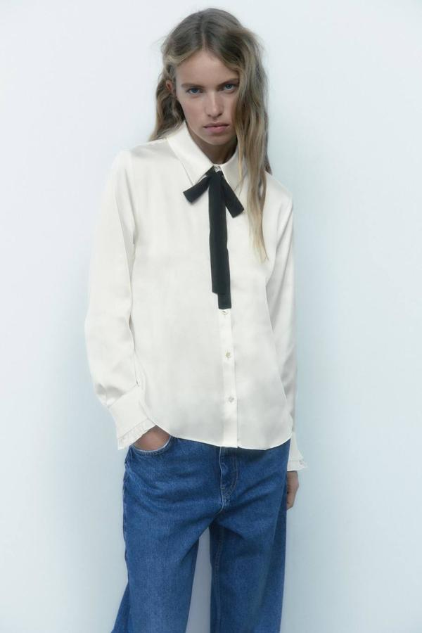 Camisa satinada blanca con cuello solapa, manga larga, cierre de botones delantero y detalle de lazo negro a contraste, de Zara. Su precio es de 27,95 euros.