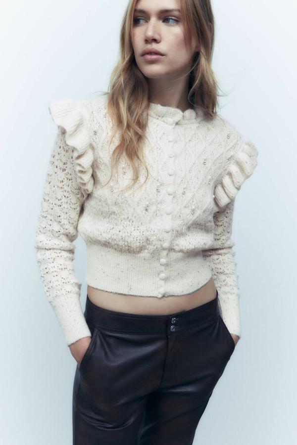 Chaqueta de punto trenzado con detalles calados, volantes, cuello redondo, terminaciones festoneadas, manga larga y aplique de cristales y perlas, en crudo. La encontrarás por 35,95 euros.