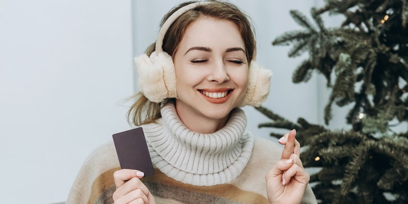 Hopeful young woman in fluffy earmuffs and striped poncho sitting in gray chair crossing fingers and holding credit card with silver colored laptop on her knees against light curtain and christmas tree with electric garland.