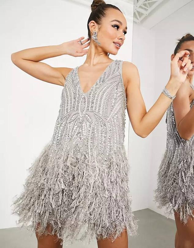 Vestido corto con aplicación de abalorios, corte recto, escote de pico, tirantes anchos y bajo acabado en plumas, color plata, de Asos. Su precio es de 175,99 euros.