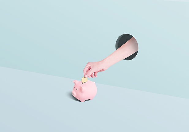 Human hand sticking through a hole in the wall placing a gold coin into a pink piggy bank on a greenish two toned background