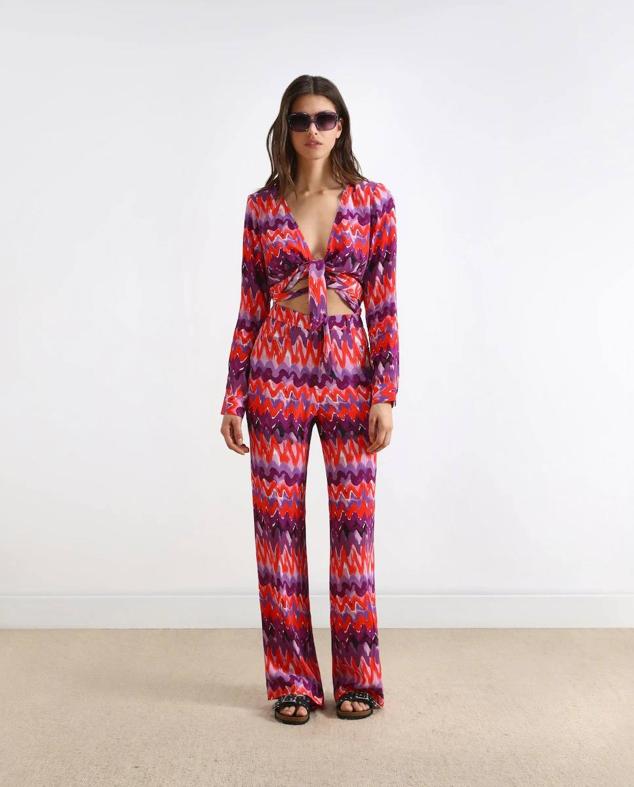 Conjunto con blusa crop con lazada y pantalón largo fluido, en estampado de zigzag morado y rojo, de Scalpers. Su precio es de 79,90 euros y 89,90 euros.