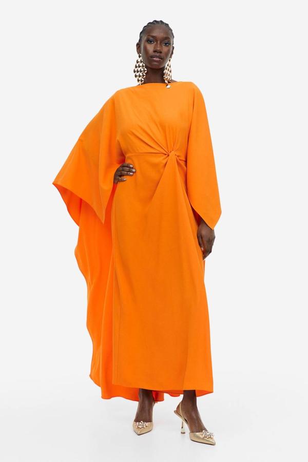 Vestido capa en tejido de crepé con escote barco, nudo decorativo en el centro y mangas larga maxi volumen, en naranja. Hazte con él por 59,99 euros.
