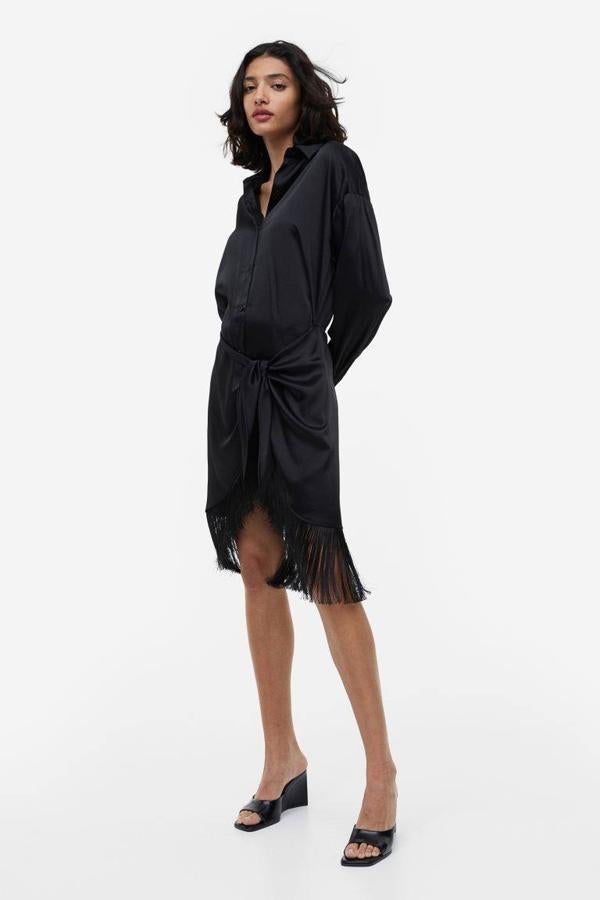 Vestido corto tipo camisero en tejido satinado con cuello solapa, manga larga, cierre de botones, detalle de nudo y bajo terminado en flecos, color negro, de H&M. Cuesta 49,99 euros.