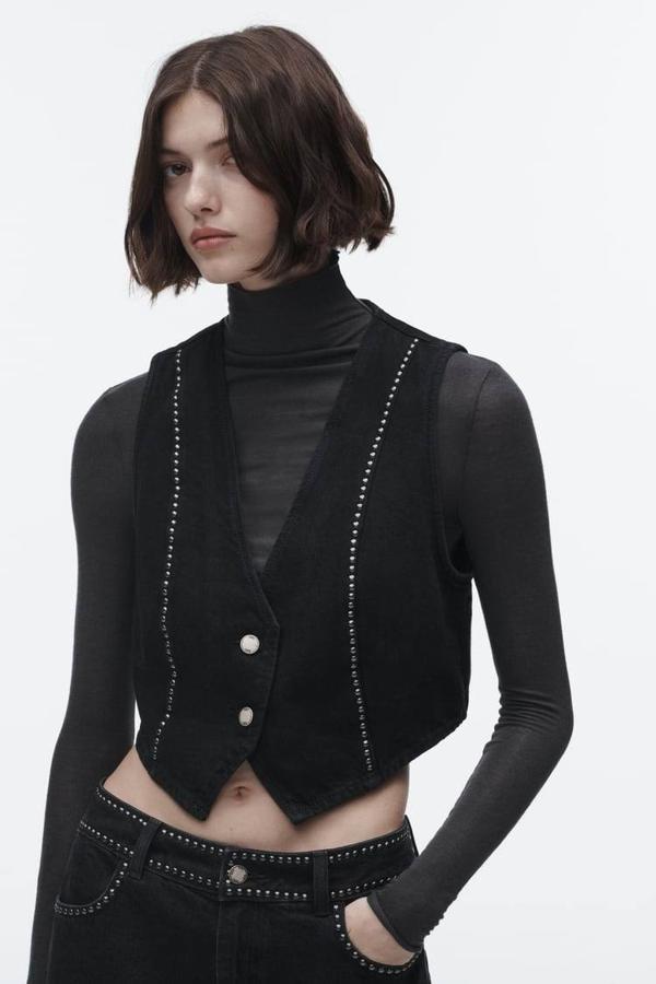 Chaleco de vestir en tejido vaquero con bajo asimétrico, manga sisa, escote de pico y detalle de tachas metálicas, color negro, de Zara. Encuéntralo por 22,95 euros.