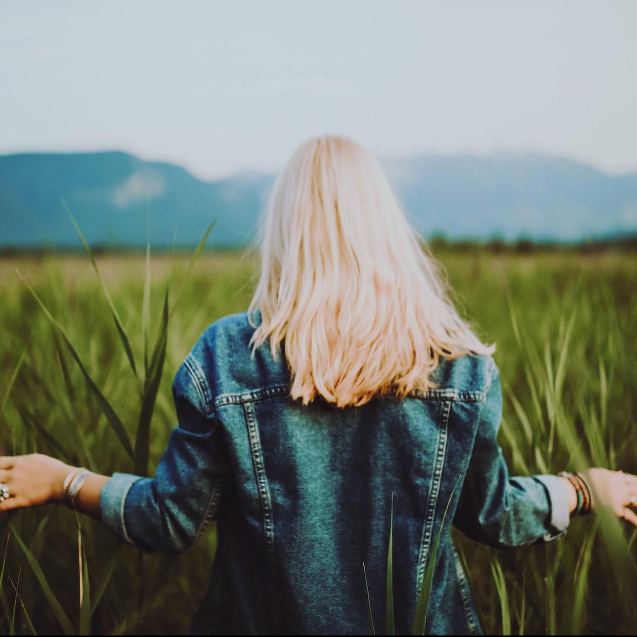 Mujer rubia con cazadora vaquera paseando por un campo verde. 