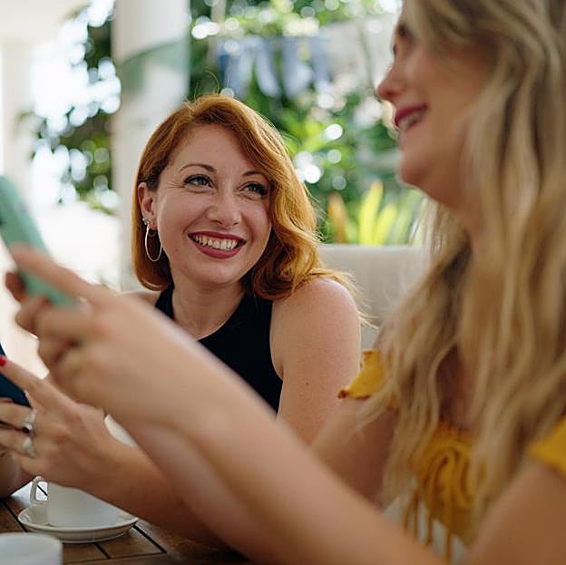 Dos mujeres conversando en una cafetería mientras miran sus móviles. 