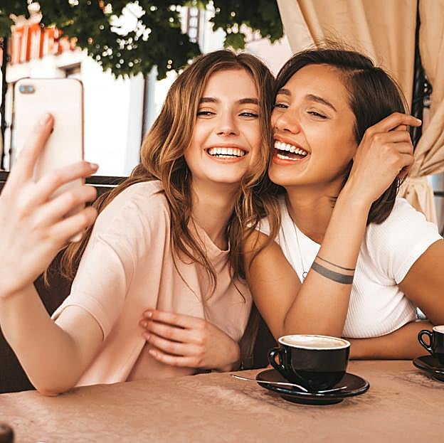 Dos amigas sonriendo haciéndose un selfie mientras toman un café. 