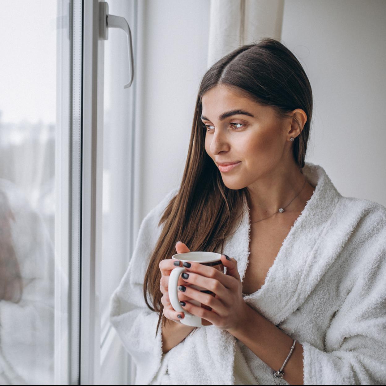 Mujer tomando una taza de infusión frente a la ventana. 
