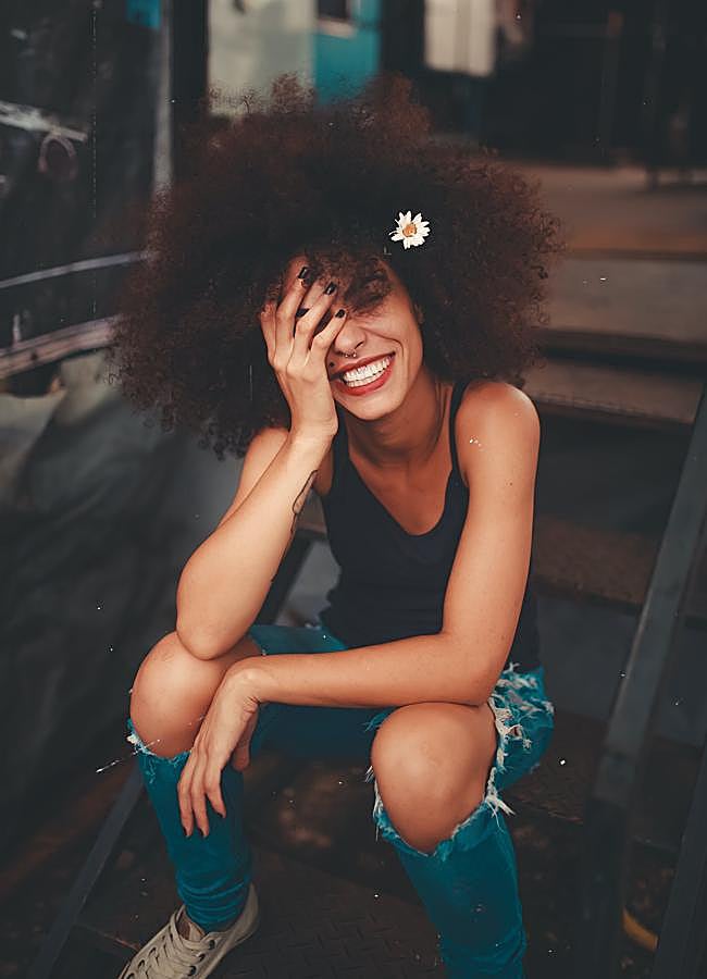 Imagen - Mujer con el pelo afro sonriendo y sentada en unas escaleras. / Foto de luizclas en Pexels. 