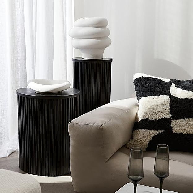 Imagen principal - Arriba, mesa auxiliar en madera acanalada en negro (también disponible en tono natural y en blanco), 159 euros. Abajo, mesa de silueta curva que combina mármol y madera en tono wengué (229 euros) y side table con diseño cúbico y efecto mármol en color gris (también disponible en blanco), 199 euros.