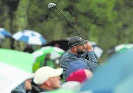 Jon Rahm golpea la bola desde el tee rodeado de aficionados con paraguas para protegerse de la lluvia.