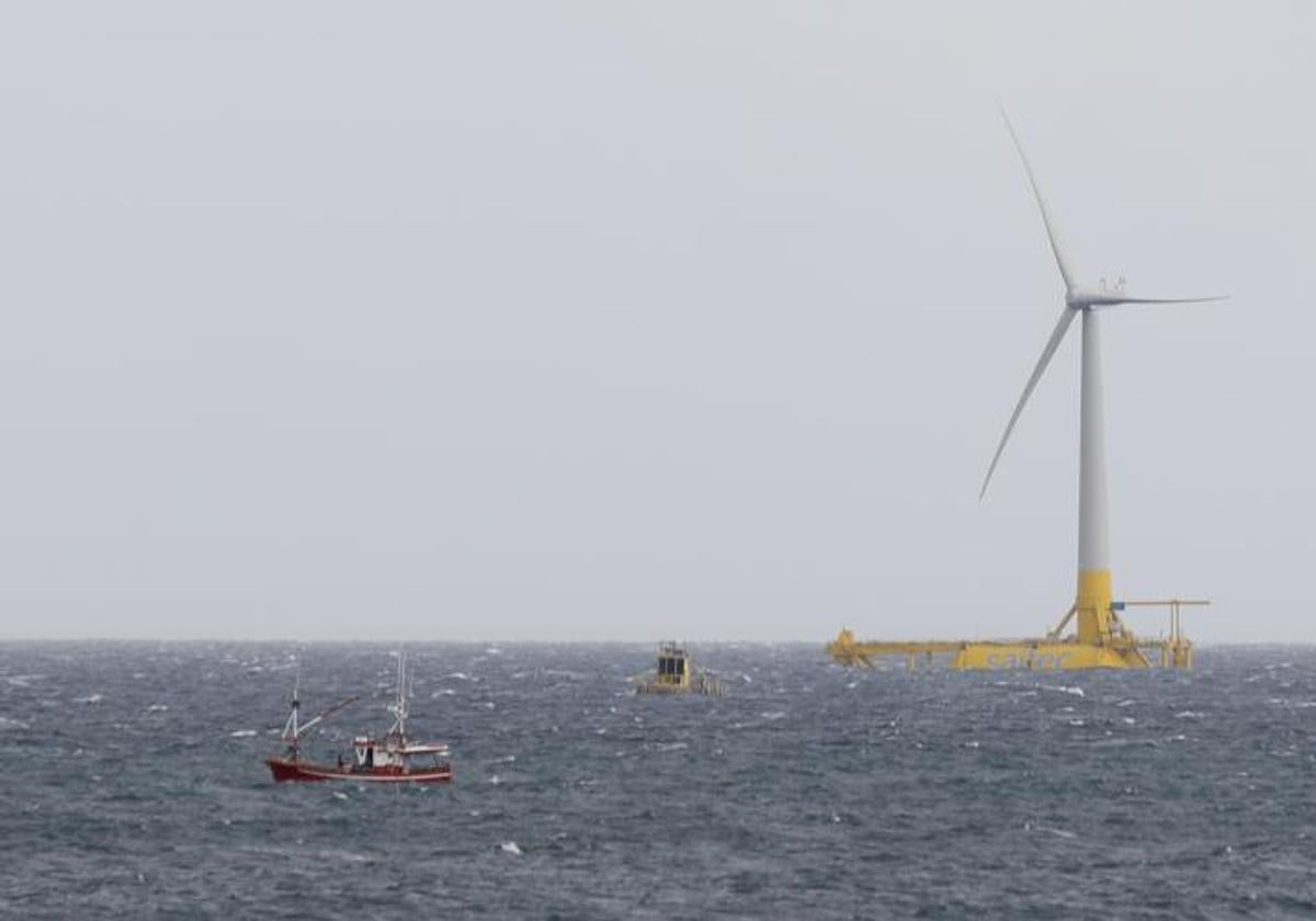 Un barco pesquero junto al prototipo (fondo) de aerogenerador de eólica marina flotante, anclado con cadenas al fondo marino .