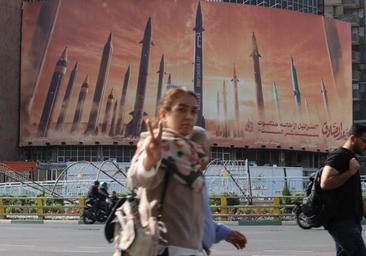 Una iraní hace la señal de la victoria mientras pasa frente a un cartel que arremete contra Israel en las calles de Teherán.