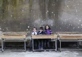 Niños de Alepo sentados en el pupitre de una escuela en cuya pared se aprecian impactos de bala.