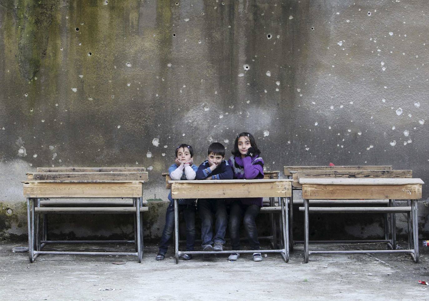 Niños de Alepo sentados en el pupitre de una escuela en cuya pared se aprecian impactos de bala.