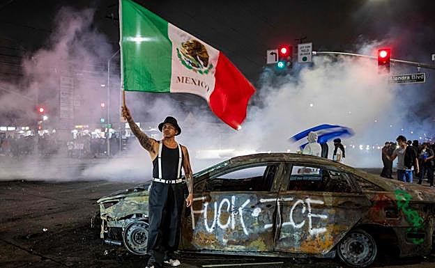 Un joven alza una bandera mexicana junto a un coche quemado.
