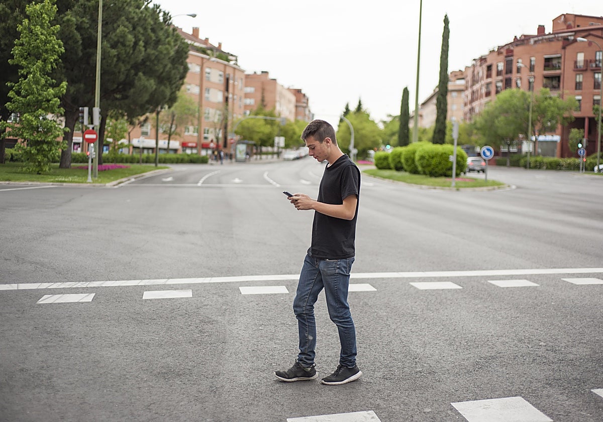 Un joven cruza una calle con la vista puesta en el móvil.