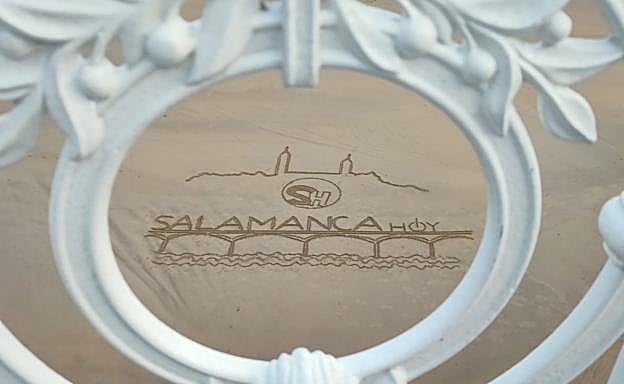 El logotipo de 'Salamancahoy' grabado en la arena de playa visto a través de la emblemática barandilla de La Concha de San Sebastián.