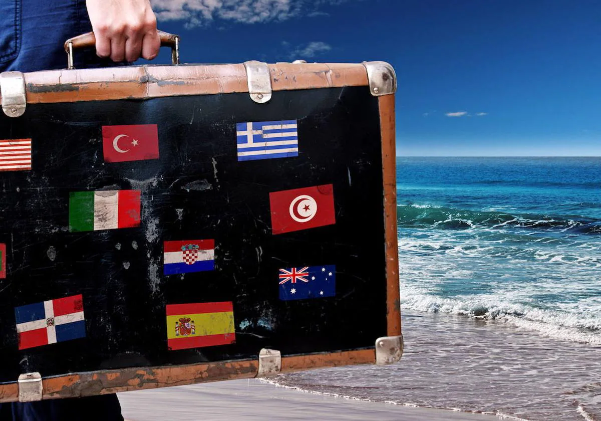 Una maleta de viaje con banderas de distintos países junto a la orilla del mar.