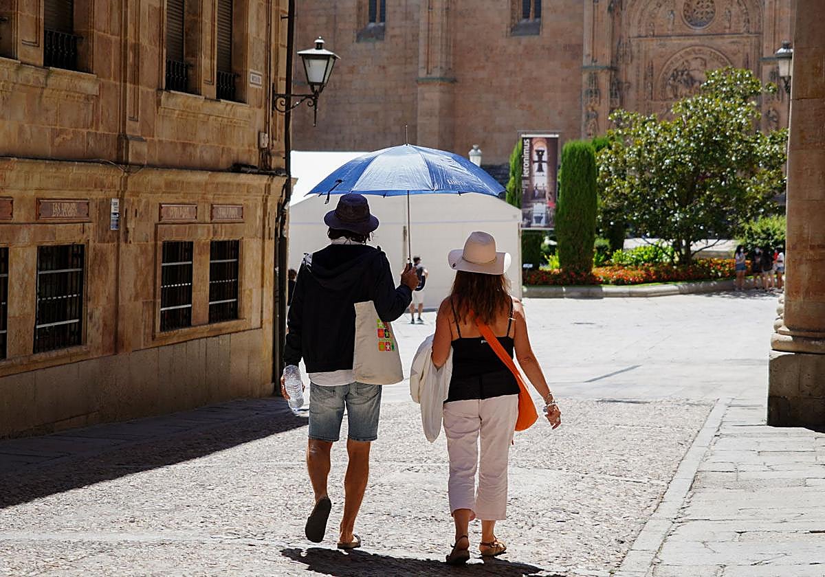 Dos turistas pasean hacia la catedral de Salamanca protegidos con sombreros y paraguas por el sol.